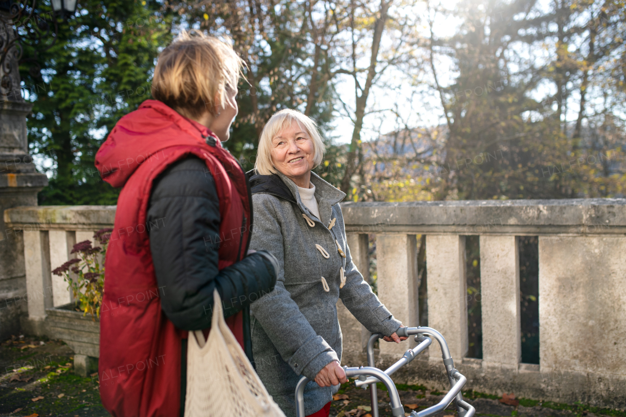 Senior woman with walking frame and caregiver or healthcare worker outdoors on a walk in park.