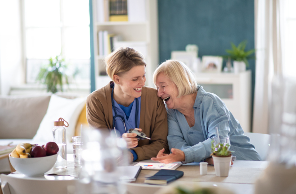 Healthcare worker with cheerful laughing senior woman patient, measuring blood glucose indoo