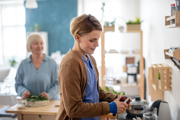 Happy senior woman with caregiver or healthcare worker indoors, preparing food.