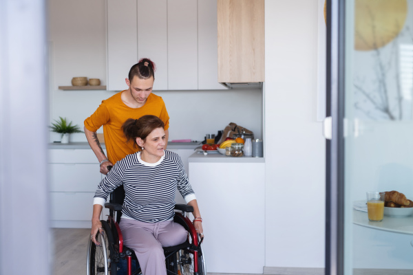 An adult son helping disabled mature mother in wheelchair in kitchen indoors at home.