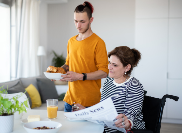 Portrait of disabled mature woman in wheelchair sitting at the table with a son indoors at home, eating.