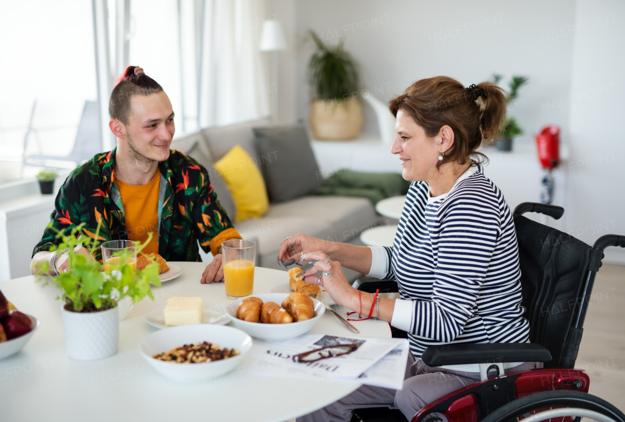 Portrait of disabled mature woman in wheelchair sitting at the table with a son indoors at home, eating.