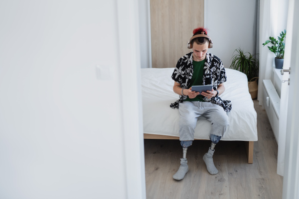 A portrait of disabled young man using tablet on bed indoors at home, leg prosthetic concept.