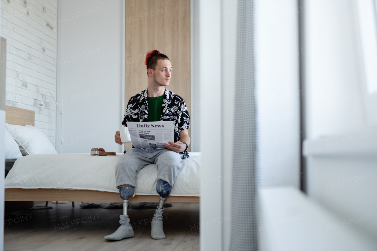 A portrait of disabled young man reading newspapers on bed indoors at home, leg prosthetic concept.