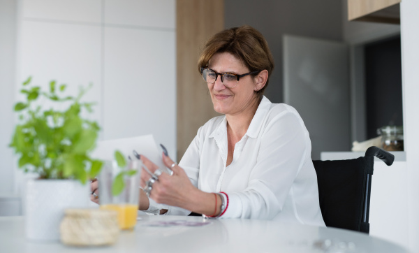 Portrait of happy disabled mature woman in wheelchair indoors at home, using tablet.