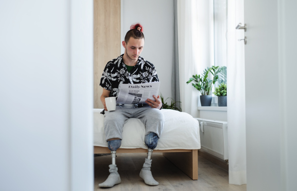 A portrait of disabled young man reading newspapers on bed indoors at home, leg prosthetic concept.