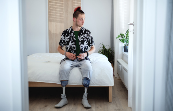 Front view portrait of disabled young man sitting in bed indoors at home, leg prosthetic concept.