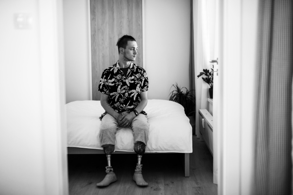 A black and white portrait of disabled young man sitting in bed indoors at home, leg prosthetic concept.