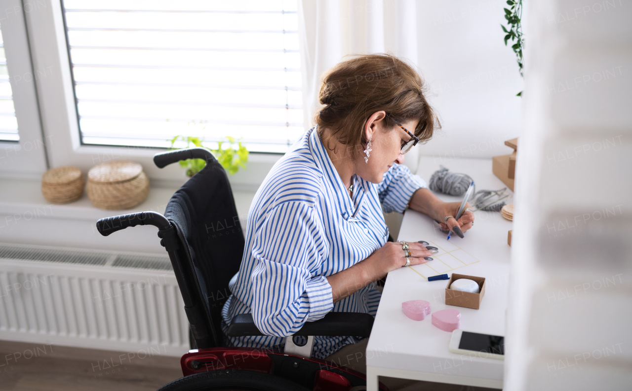 A disabled mature woman in wheelchair packing handmade products indoors at home, small business concept.