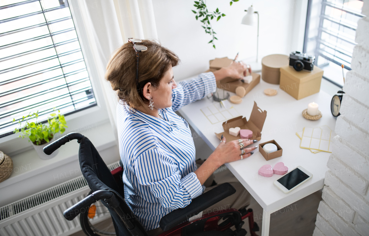 A disabled mature woman in wheelchair packing handmade products indoors at home, small business concept.