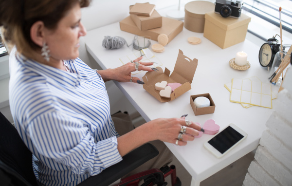 A disabled mature woman in wheelchair packing handmade products indoors at home, small business concept.