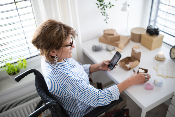 A disabled mature woman in wheelchair packing handmade products indoors at home, small business concept.