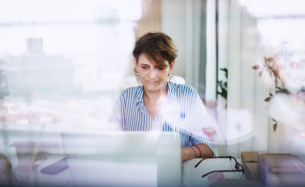 A mature woman with laptop working indoors in home office, small business concept. Shot through glass.