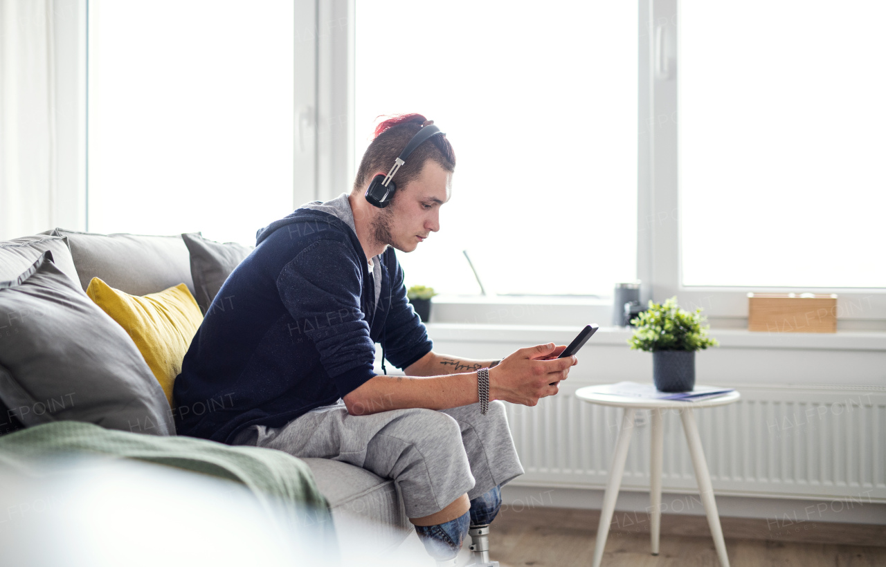 A portrait of disabled young man using smartphone indoors at home, leg prosthetic concept.