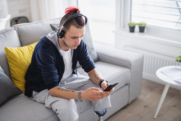 A portrait of disabled young man using smartphone indoors at home, leg prosthetic concept.