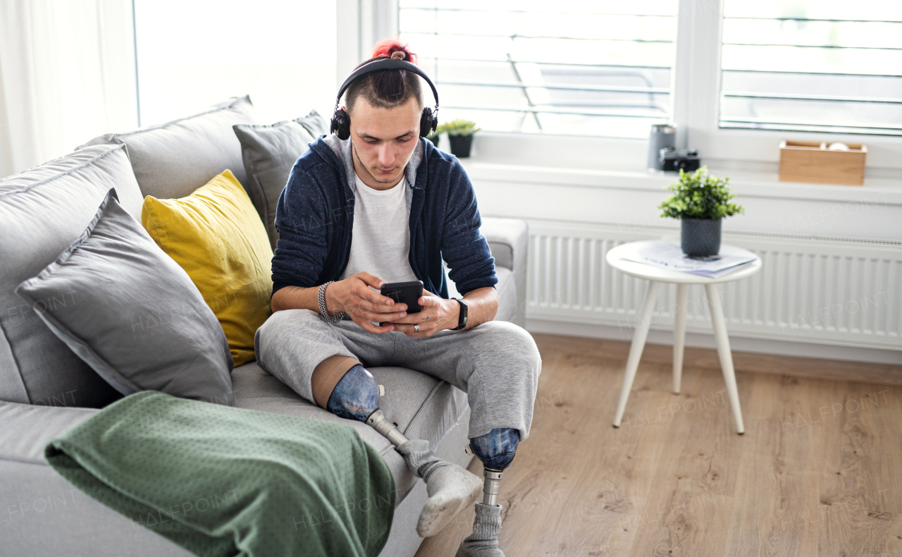 A portrait of disabled young man using smartphone indoors at home, leg prosthetic concept.