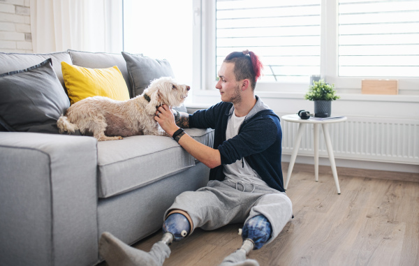 A portrait of disabled young man playing with dog indoors at home, leg prosthetic concept.