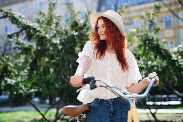 A portrait of young woman commuter with bicycle outdoors in city in summer.