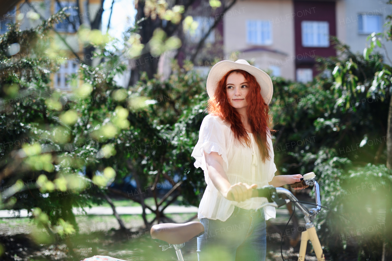 A portrait of young woman commuter with bicycle outdoors in city in summer.