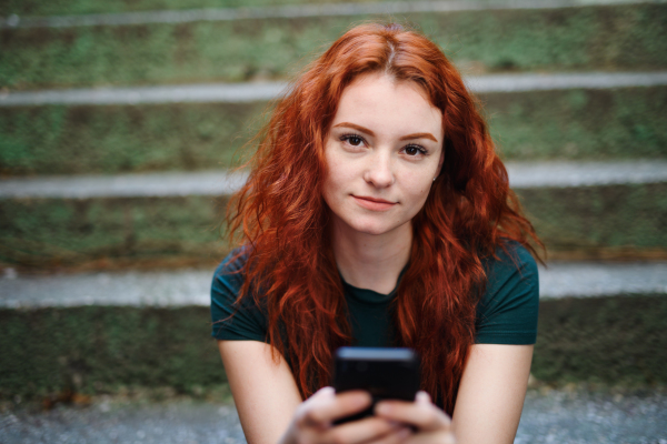 A portrait of young woman with smartphone outdoors in city street, looking at camera.