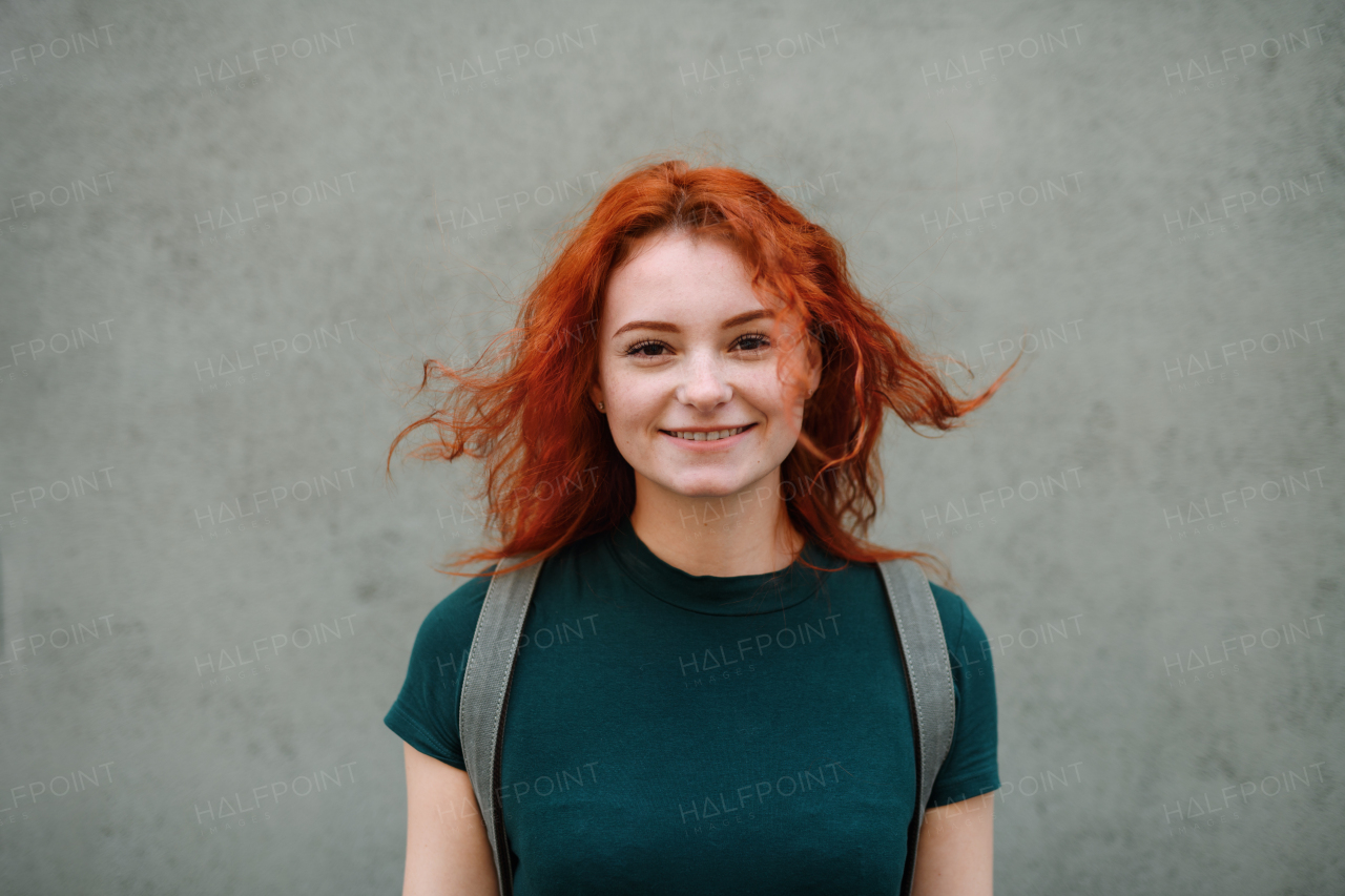 A portrait of young woman standing outdoors against gray background, looking at camera.