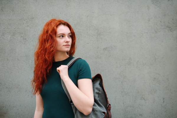 A portrait of young woman with backpack standing outdoors against gray background. Copy space.