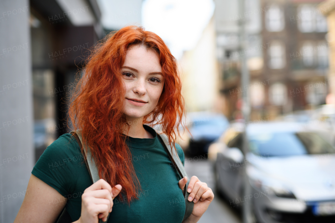 A portrait of young woman with backpack standing outdoors in city, looking at camera.