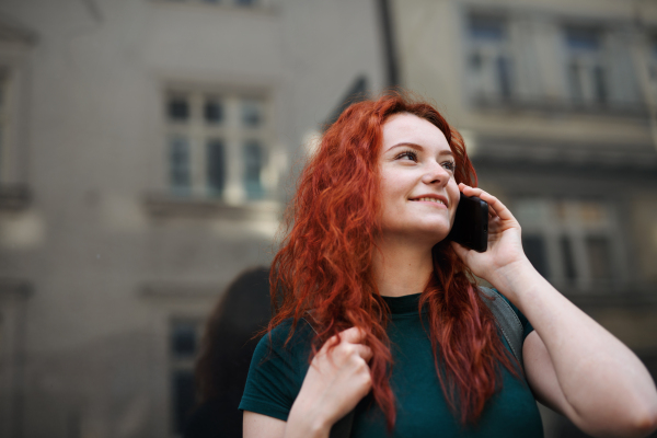 A portrait of young woman with backpack standing outdoors on city street, using smartphone.