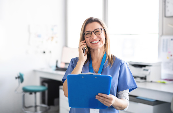 A portrait of cheerful dental assistant in modern dental surgery, using smartphone.