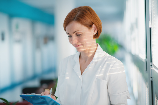 Portrait of woman doctor with clipboard standing in hospital. Shot through glass.