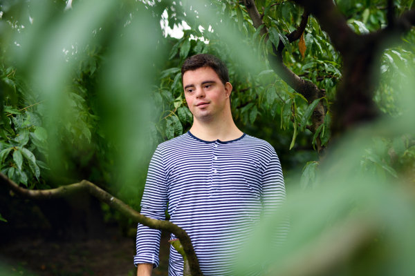 Front view portrait of down syndrome adult man standing outdoors under tree.