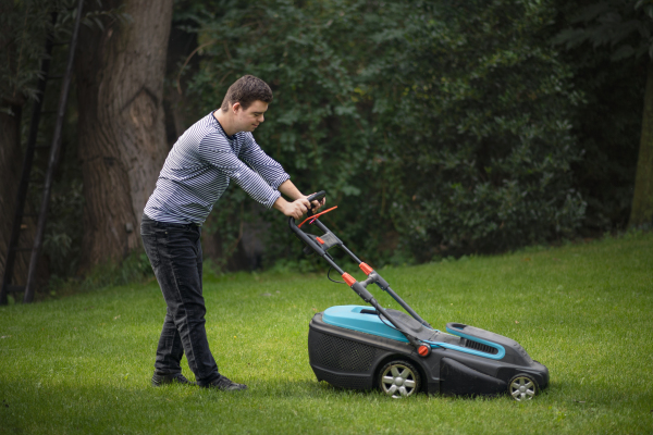 Portrait of down syndrome adult man mowing lawn outdoors in backyard, helping with housework concept.