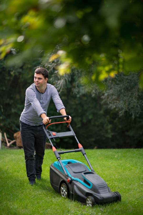 Portrait of down syndrome adult man mowing lawn outdoors in backyard, helping with housework concept.