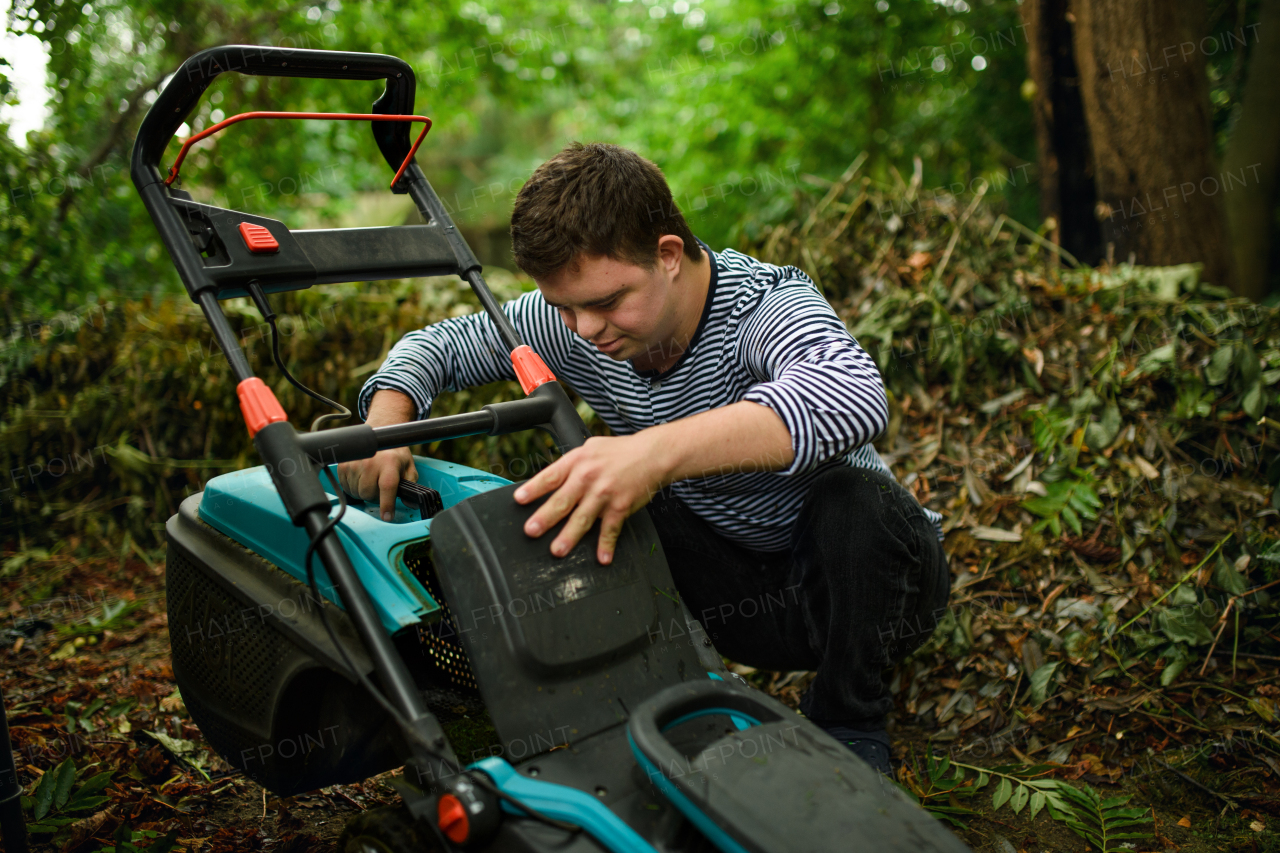 Down syndrome adult man emptying grass box in lawn mower outdoors in backyard, helping with housework concept.