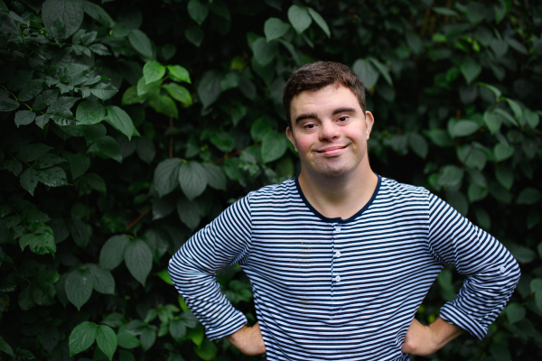 Portrait of down syndrome adult man standing outdoors at green leaves background, looking at camera.