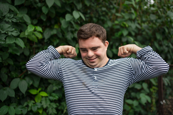 Portrait of down syndrome adult man standing outdoors at green leaves background, flexing muscles.