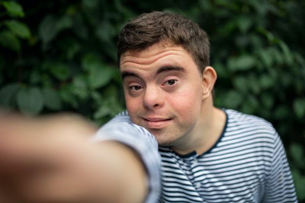 Front view portrait of down syndrome adult man standing outdoors in garden, taking selfie.