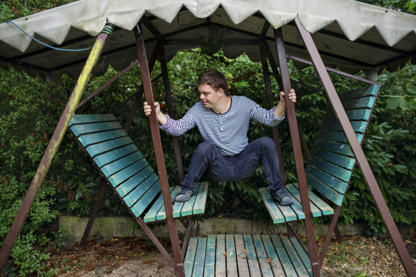 Front view portrait of down syndrome adult man outdoors on swing in garden.