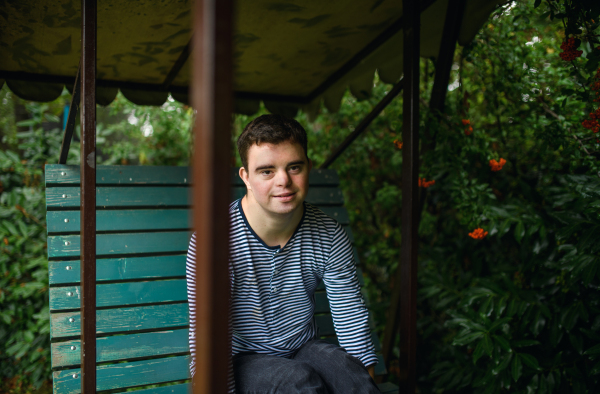 A portrait of down syndrome adult man sitting outdoors in garden, looking at camera.