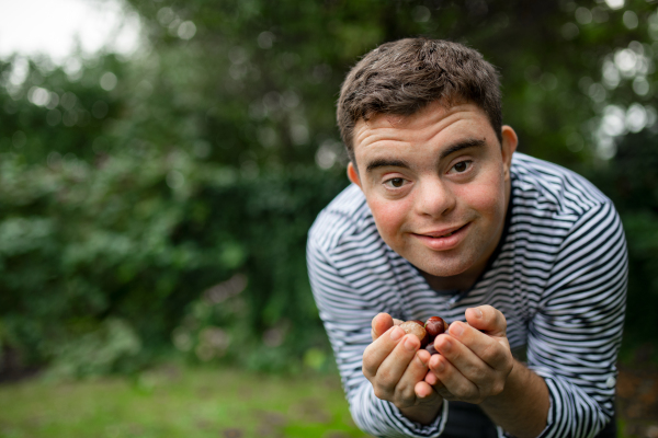 Portrait of down syndrome adult man standing outdoors at green background, holding conkers in hands.