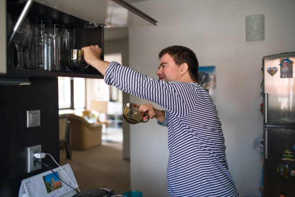 Portrait of down syndrome adult man standing indoors in kitchen at home, helping with dishes.