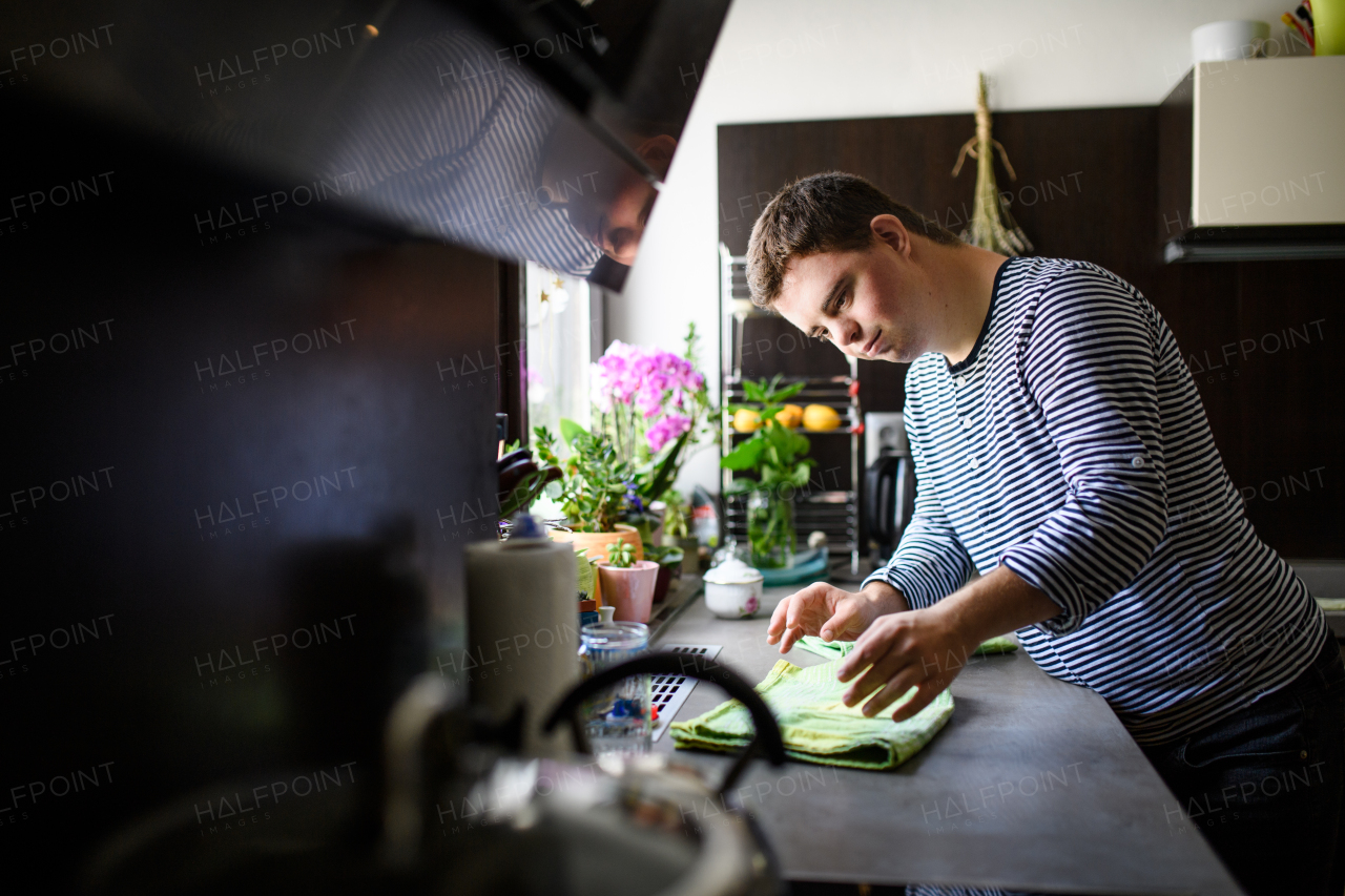 Portrait of down syndrome adult man standing indoors in kitchen at home, helping.