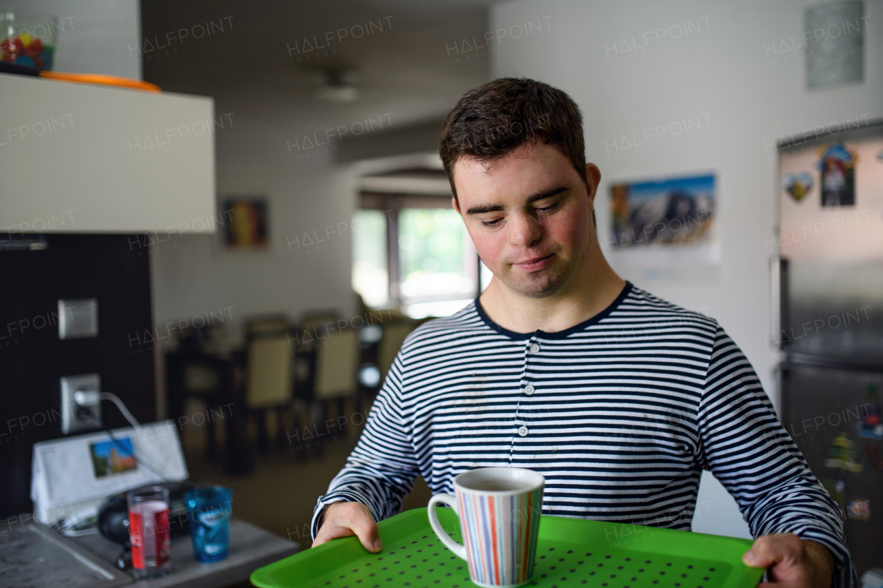 Portrait of down syndrome adult man standing indoors in kitchen at home, carrying cup on tray.