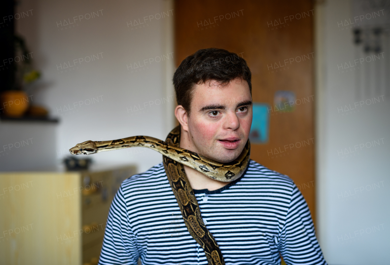 Portrait of down syndrome adult man sitting indoors in bedroom at home, playing with pet snake.