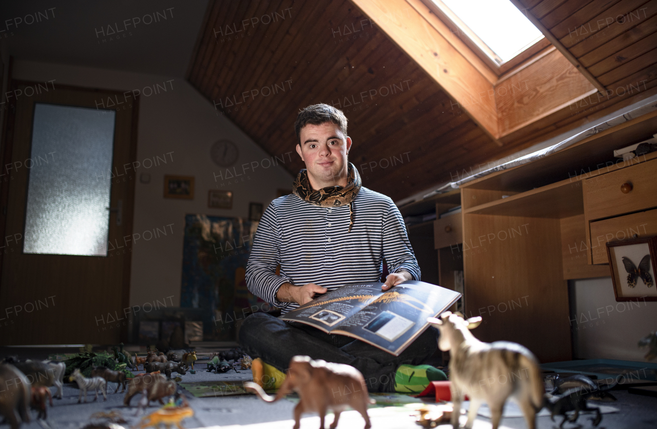 Portrait of down syndrome adult man with pet snake sitting indoors in bedroom at home.