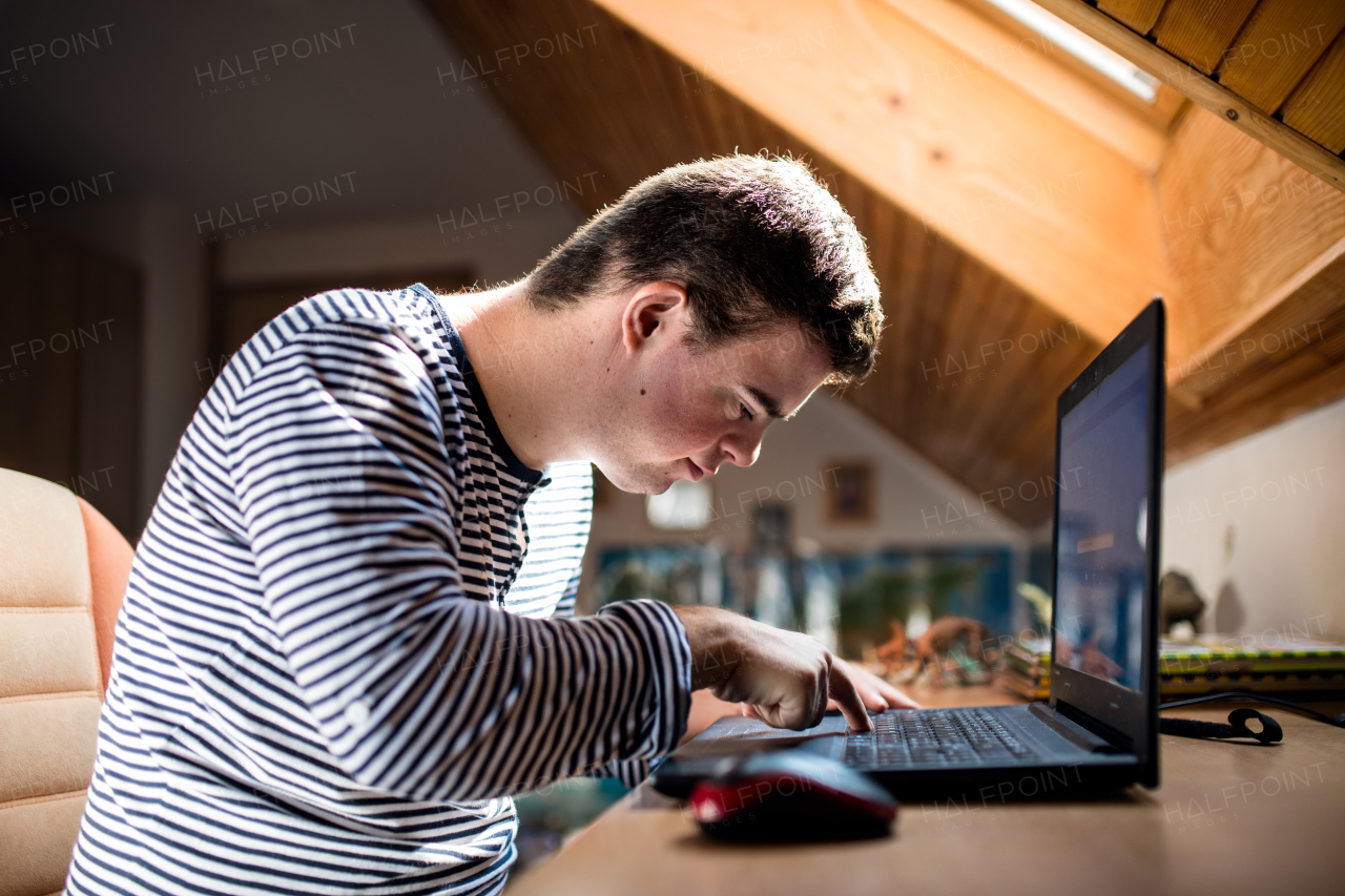 Happy down syndrome adult man sitting indoors in bedroom at home, using laptop.