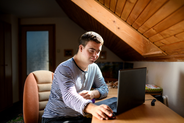 Happy down syndrome adult man sitting indoors in bedroom at home, using laptop.