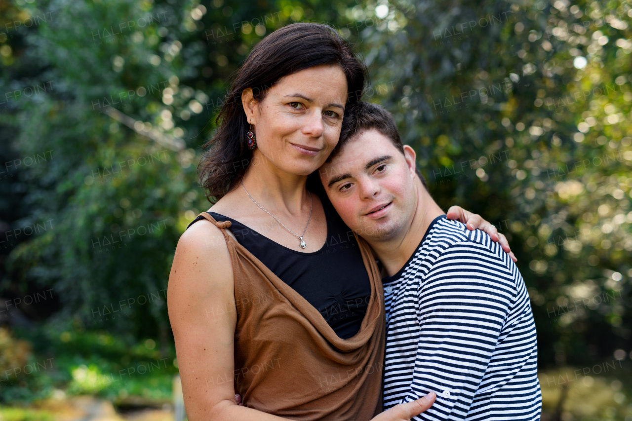 Portrait of down syndrome adult man with mother standing outdoors in garden, looking at camera.