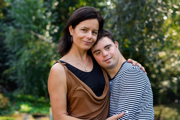 Portrait of down syndrome adult man with mother standing outdoors in garden, looking at camera.