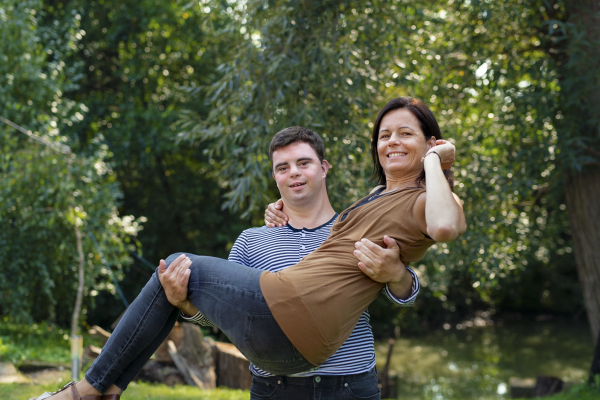 Portrait of happy down syndrome adult man carrying mother in arms outdoors in garden.